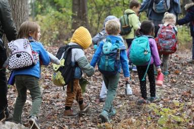 Preschool children wearing backpacks walk in a line holding hands through an autumn forest.