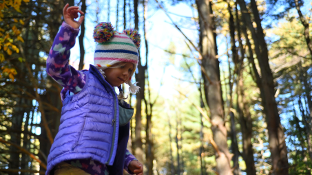 A young girl wearing a puffy purple vest and a knit cap in a forest.