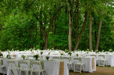 Rows of white tables and chairs set up for a fancy dinner in a clearing surrounded by a forest.