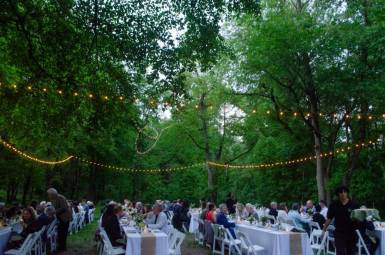 Rows of white tables set for dinner in a clearing surrounded by tall green trees.