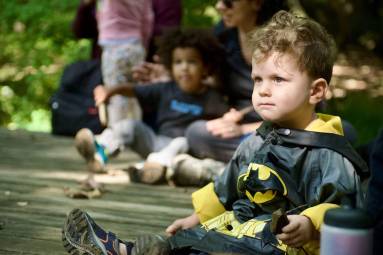 A toddler dressed in a batman costume sits on a wood platform while children and parents sit and stand in the backround.