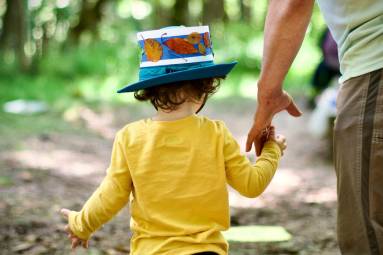 A child in a yellow shirt and blue hat with autumn leaves holds the hand of an adult on a train in a green forest.