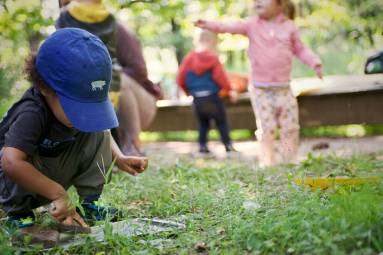 A child in a t-shirt and blue hat crouches looking at the ground while other small children play behind him in a forest.