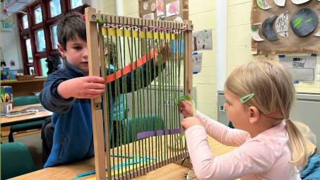 Two preschool children play with a loom in an decorated classroom with large windows.