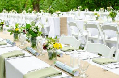 A white table with white chairs decorated with flowers and with place settings for dinner in a outdoor natural setting.