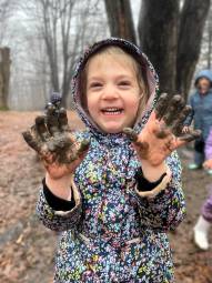 A young child i n rain gear smiles while showing off muddy hands.