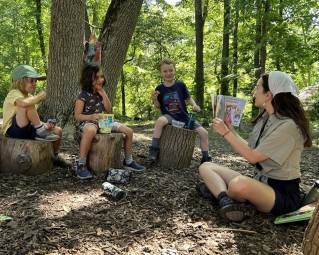 A teacher reads aloud to three preschool children sitting on stumps outdoors.