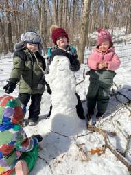 Four children in winter clothing surround a snowman in snowy forested landscape.