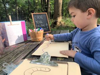 A young child draws an earthworm on a wooden table outside.