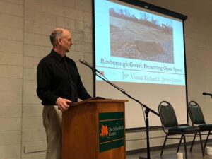 A man speaking at a podium with a power point presentation on display to the side of him titled Roxborough Green Preserving Open Space. 