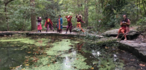 Five women sit, stand and dance next to a pond in the forest.