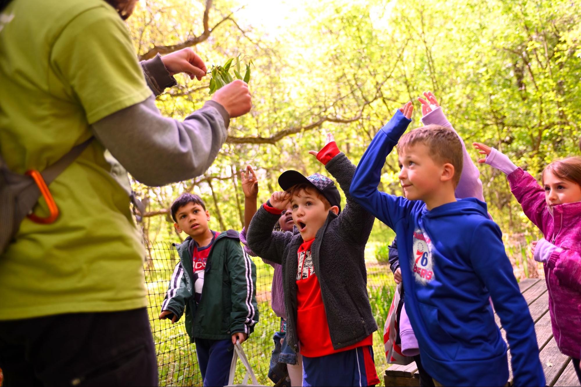 Enthusiastic children in fall weather clothes raise their hands while looking at an adult holding a plant.