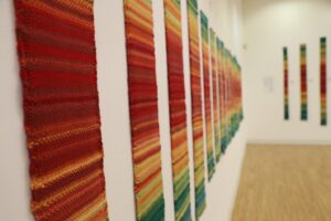 Woven scarf tapestries in red, yellow, blue, and green hanging on white walls, the colors corresponding to the temperature of the day during a year.