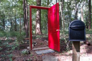 A mailbox and a red door leading to a fenced off area in a forest.