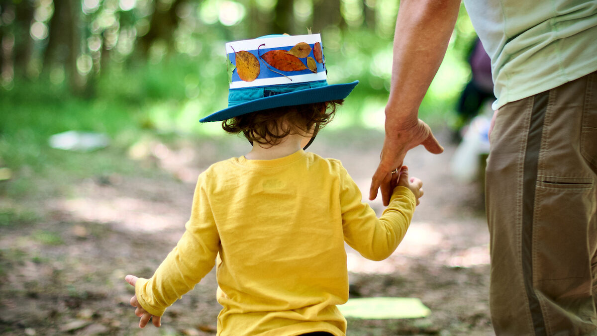 A child in a yellow shirt and blue hat with autumn leaves holds the hand of an adult on a train in a green forest.