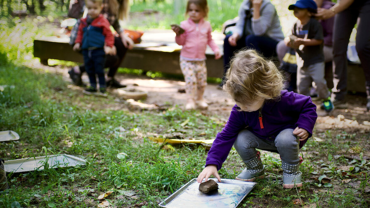 A child in a blue jacket crouches down to play while other children play in the background.