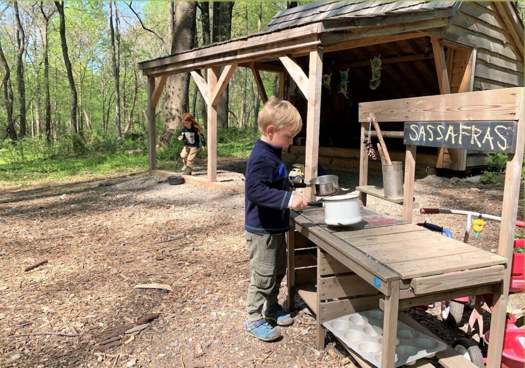 Two children play in a natural setting, one next to a small table with a chalk sign that says Sassafras and the other leaps in the background excitedly.