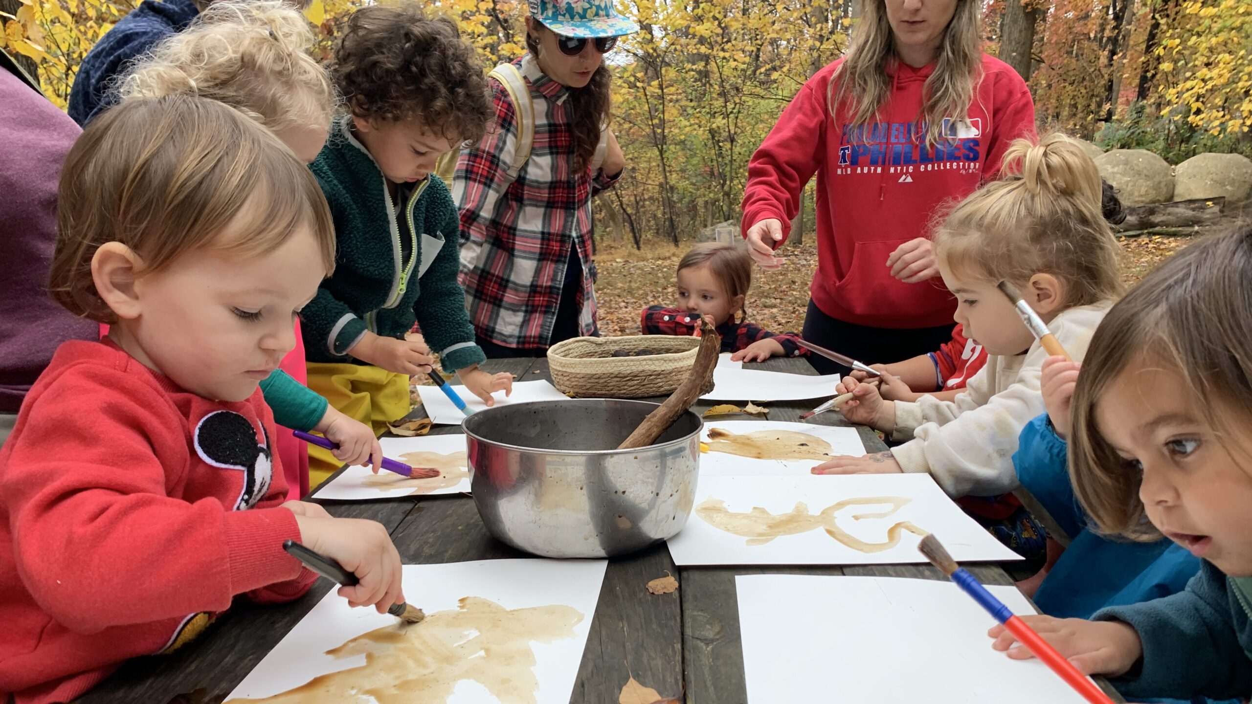 Small children sit around a wooden table outdoors making art in the autumn.