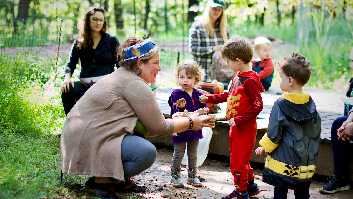 An adult wearing a crown of paper an autumn leaves crouches to talk to children in an outdoor setting.