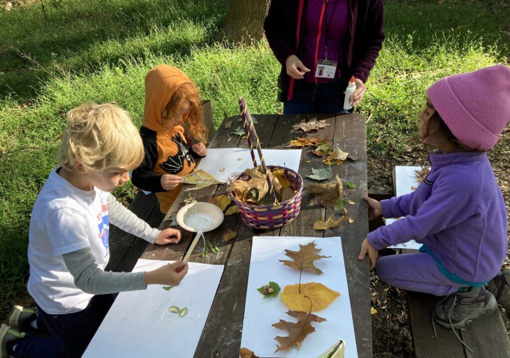 Three children paint on paper sitting at a wooden table outdoors.